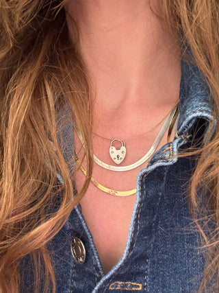 Heart padlock charm styled among layered necklaces worn at the neckline.