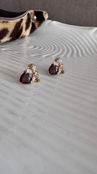 Pair of heart-shaped garnet earrings with diamond bow tops on a white surface.