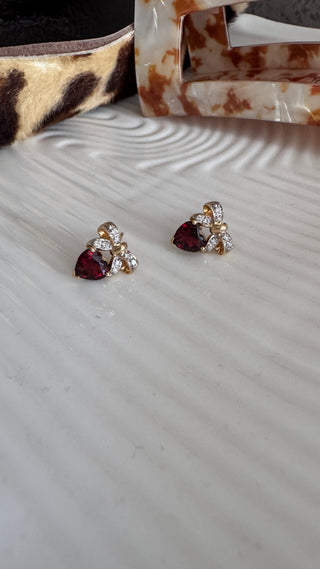 Pair of heart-shaped garnet earrings with diamond bow tops on a white surface.