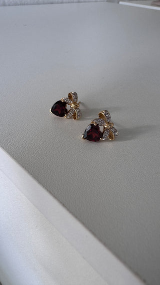 Pair of heart-shaped garnet earrings with diamond bow tops on a white surface.
