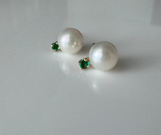 14k gold cultured pearl stud earrings with small green emerald accents on a gray surface.