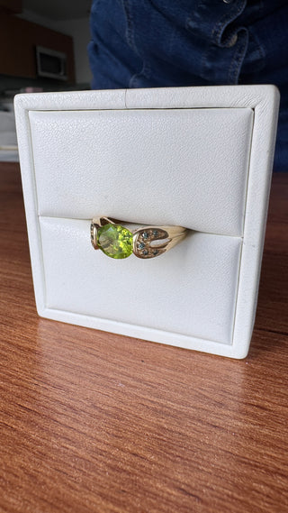 Gold peridot ring with split shoulders and blue accents in a white jewelry box.