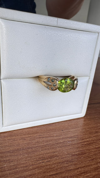 Gold peridot ring with split shoulders and blue accents in a white jewelry box.