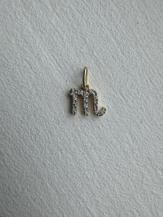 Diamond-set Scorpio symbol pendant with a gold bail on pale textured fabric.