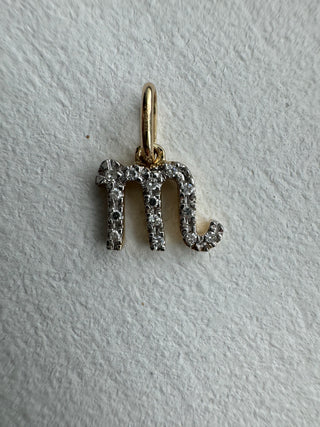 Diamond-set Scorpio symbol pendant with a gold bail on pale textured fabric.