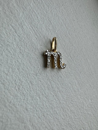 Diamond-set Scorpio symbol pendant with a gold bail on pale textured fabric.