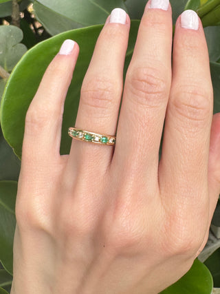 Vintage gold emerald and diamond band worn on a hand against green leaves.