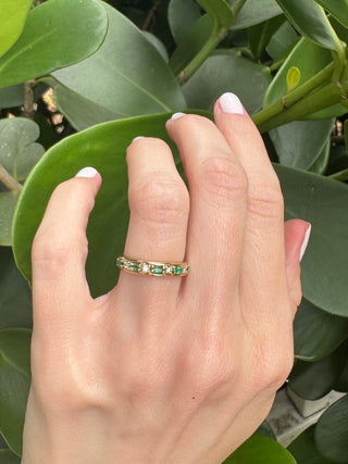 Vintage gold emerald and diamond band worn on a hand against green leaves.