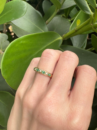 Vintage gold emerald and diamond band worn on a hand against green leaves.