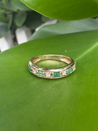 Gold band with alternating green emeralds and diamonds resting on a green leaf.