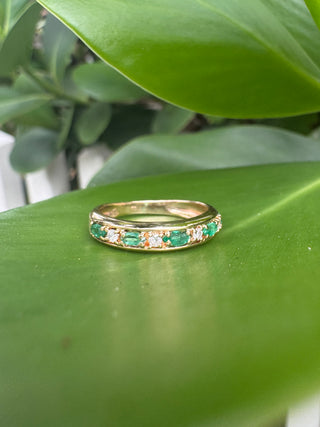 Gold band with alternating green emeralds and diamonds resting on a green leaf.