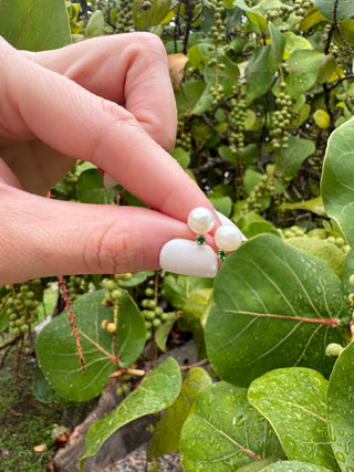 Cultured pearl and emerald stud earrings held between fingers against green leaves.