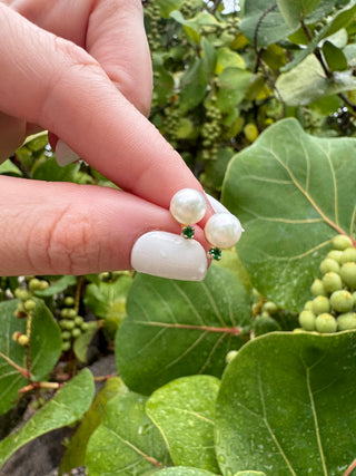 Cultured pearl and emerald stud earrings held between fingers against green leaves.