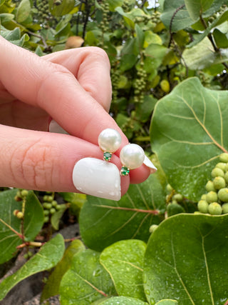 Cultured pearl and emerald stud earrings held between fingers against green leaves.