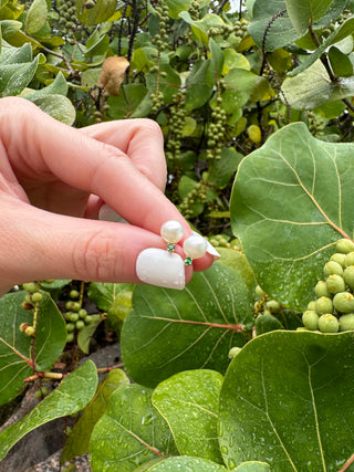 Cultured pearl and emerald stud earrings held between fingers against green leaves.