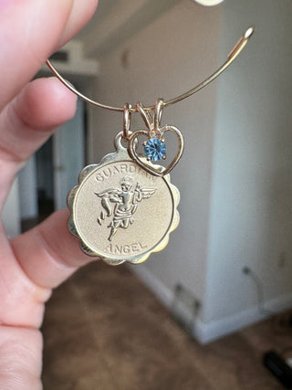 14k gold aquamarine heart charm hanging beside a larger zodiac pendant, held in a hand.