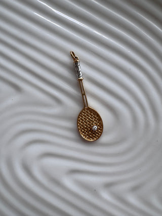 Gold tennis racket pendant on a textured white surface.