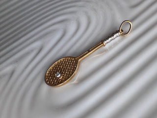 A tennis racket-shaped pendant charm in gold-tone metal with a textured head, a silver-tone wrapped handle, and a small clear stone set in the center, resting on a white wavy surface.