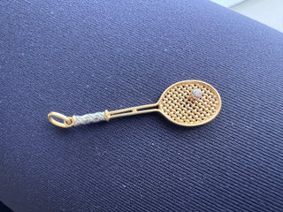 A gold tennis racket pendant with a tennis ball detail, displayed on a textured navy blue fabric background.