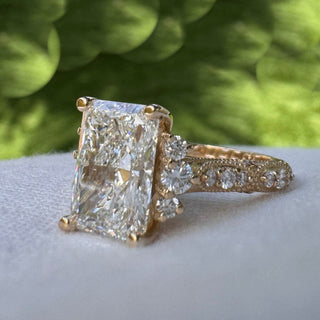 A yellow gold ring with a large rectangular clear gemstone and smaller side stones, resting on a light gray surface with a blurred outdoor garden background.